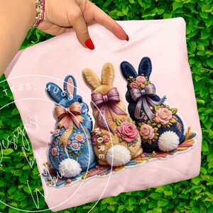 May include: A pink cushion featuring three decorative Easter bunnies. The bunnies are blue, tan, and navy, each adorned with floral accents, bows, and fluffy white tails. The cushion is held by a hand with red nail varnish.