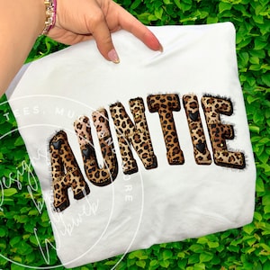 Puede incluir: Camiseta blanca con la palabra "AUNTIE" en estampado de leopardo. Las letras están delineadas en negro y tienen pequeños detalles de corazón negro. La camiseta es sostenida por una mano.
