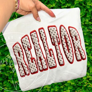 May include: White t-shirt with the word "REALTOR" in large, bold letters. The letters are white with black polka dots and outlined in red. The shirt is held up by a hand.