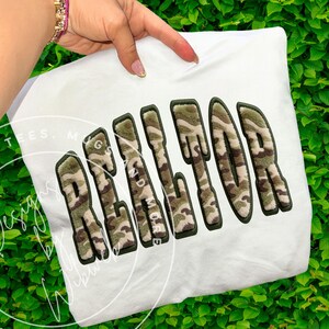 May include: A white sweatshirt featuring the word "REALTOR" in large, textured letters. The letters have a camouflage pattern with a dark green outline. The words "AND MORE" are below the word "REALTOR".