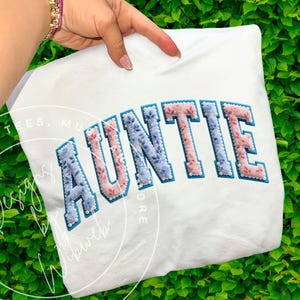 Puede incluir: Sudadera blanca con la palabra "AUNTIE" bordada con flores azules y rosas. Las letras están delineadas en un azul más oscuro y acentuadas con pequeñas cuentas blancas. Una mano sostiene la sudadera.