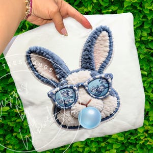 May include: White cushion featuring a blue and white embroidered bunny wearing sunglasses and a blue bubblegum bubble. The sunglasses have a floral pattern. The text "TEES MUGS AND" is visible.