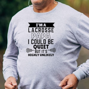 Op de afbeelding: Grijs longsleeve shirt met zwarte tekst die luidt "Ik ben een lacrosse-papa, ik zou stil kunnen zijn, maar het is zeer onwaarschijnlijk". De tekst wordt omringd door een zwarte banner en twee lacrosse-sticks.