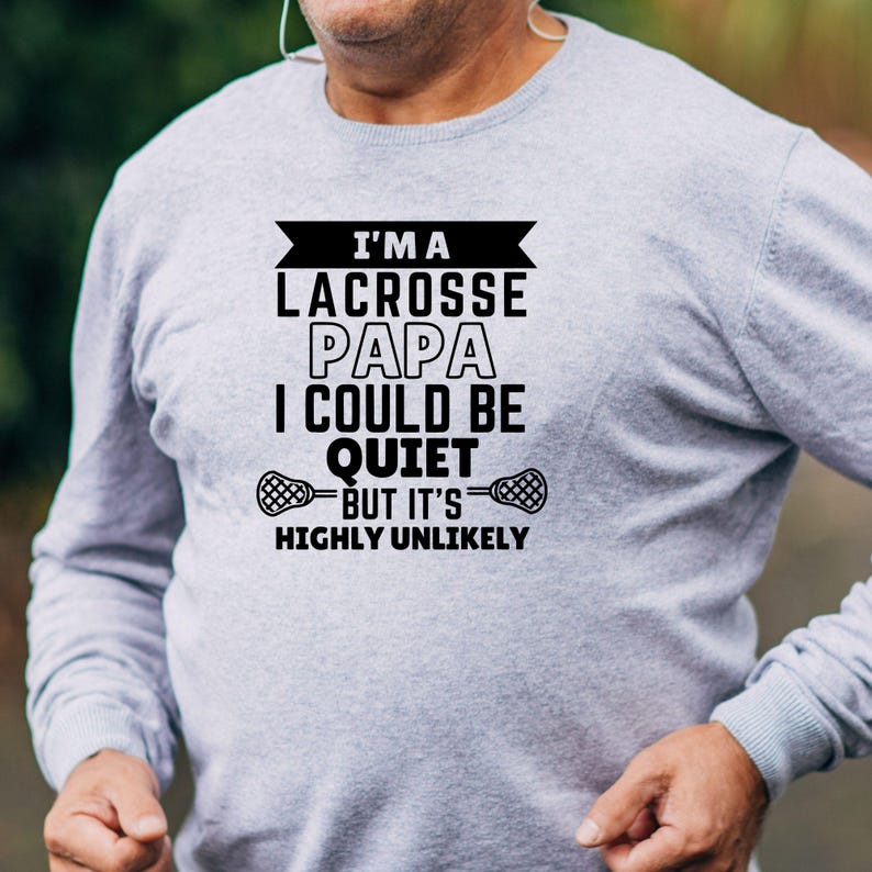 Op de afbeelding: Grijs longsleeve shirt met zwarte tekst die luidt "Ik ben een lacrosse-papa, ik zou stil kunnen zijn, maar het is zeer onwaarschijnlijk". De tekst wordt omringd door een zwarte banner en twee lacrosse-sticks.