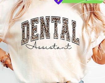 May include: Cream-colored t-shirt with the words "DENTAL Assistant" in a bold, leopard print design. The text is arched across the front, with the word "Assistant" in a cursive font below. A small graphic is in the upper right corner.