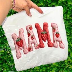 Può includere: T-shirt bianco con la scritta "NAMA" in un motivo a fragola. Le lettere sono rosa e rosse, con fragole ricamate. La maglietta è tenuta su uno sfondo verde e foglioso.