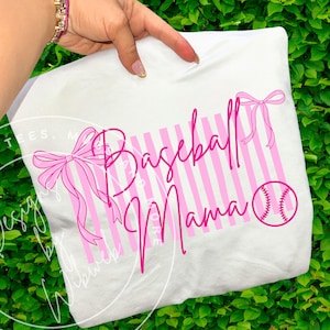 Op de afbeelding: Wit T-shirt met een roze honkbalthema-ontwerp. Het ontwerp bevat de woorden "Baseball Mama" in een scriptlettertype, roze verticale strepen, een honkbalafbeelding en roze strikken. Het shirt wordt door een hand omhoog gehouden.