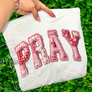 May include: White t-shirt featuring the word "PRAY" in appliquéd pink fabric letters. The letters have a patchwork design with polka dots, gingham, and floral patterns. The shirt is held by a person with a gold bracelet.
