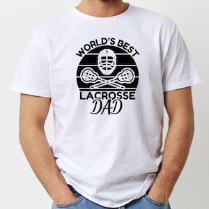 Op de afbeelding: Wit T-shirt met een zwart grafisch ontwerp met de tekst "World's Best Lacrosse Dad" met een lacrosse-stick en helm.