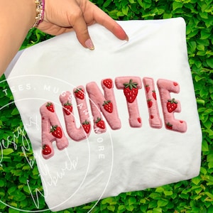 Puede incluir: Camiseta blanca con la palabra "AUNTIE" en rosa, letras decoradas con fresas. Cada letra está adornada con fresas bordadas. La camiseta está sujeta por una mano con una pulsera rosa.