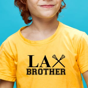 Op de afbeelding: Een geel T-shirt met zwarte tekst die "LAX BROTHER" zegt en een paar lacrosse-sticks.
