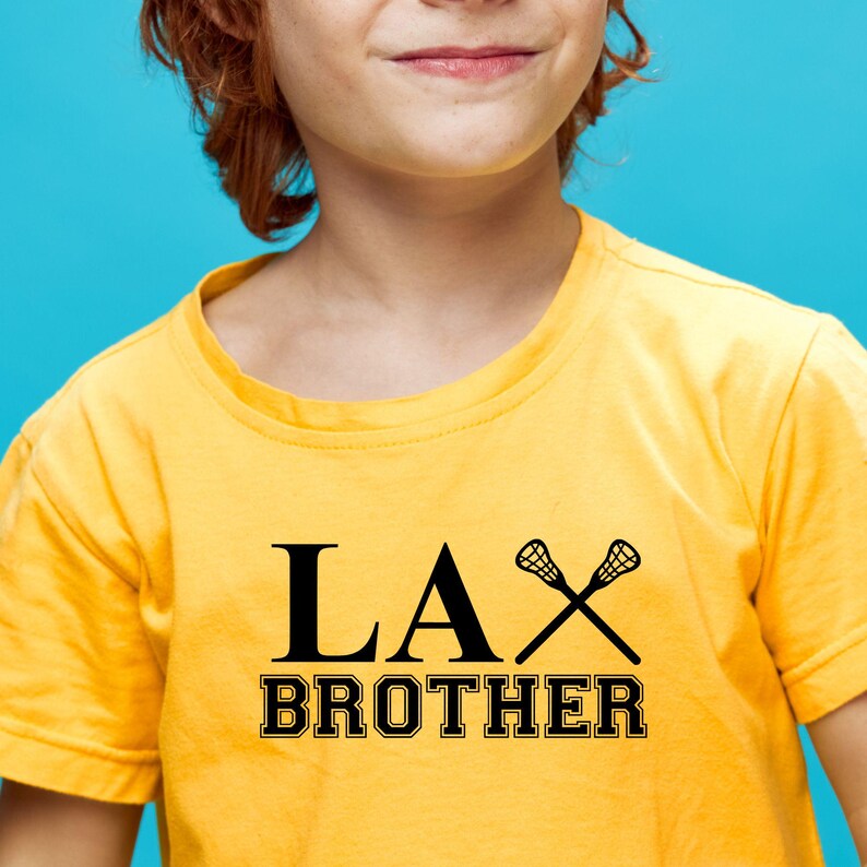 Op de afbeelding: Een geel T-shirt met zwarte tekst die "LAX BROTHER" zegt en een paar lacrosse-sticks.