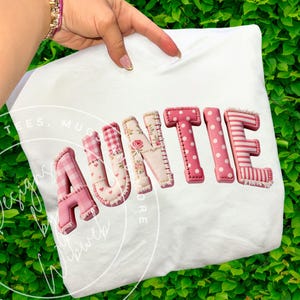 Puede incluir: Camiseta blanca con la palabra "AUNTIE" en letras rosas estampadas. Las letras tienen varios diseños, incluyendo lunares, rayas y estampados florales. La camiseta está sujeta por una mano.