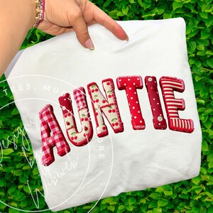 Puede incluir: Camiseta blanca con la palabra "AUNTIE" en patrones rojos, de cerezas y cuadros. Las letras están cosidas en la camiseta. La camiseta se sostiene sobre un fondo de follaje verde.