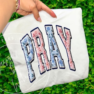 Pode incluir: Camiseta branca com a palavra "PRAY" em um design floral. As letras são contornadas em azul marinho e preenchidas com flores rosa e azuis. O design é embelezado com pequenas contas brancas.