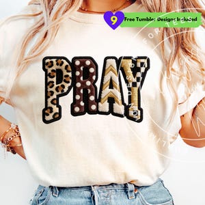 May include: Cream-colored t-shirt with the word "PRAY" in a patchwork design. The letters feature leopard print, polka dots, gold chevron, and a black and white checkered pattern. The shirt has a crew neck.