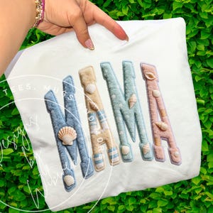 May include: White t-shirt with the word "MAMA" in pastel colors. Each letter is decorated with seashells. The letters are blue, tan, and pink, with a textured, stitched appearance. The shirt is held by a hand with a gold bracelet.