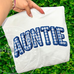 Puede incluir: Sudadera blanca con la palabra "AUNTIE" en letras grandes a cuadros azules y blancos con un borde esponjoso. La prenda se muestra sobre un fondo de follaje verde.