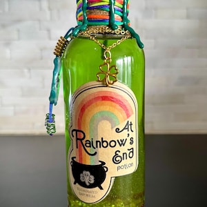 Könnte beinhalten: Eine grüne Glasflasche mit goldenem Deckel und einem Etikett mit der Aufschrift "At Rainbow's End Potion". Die Flasche ist mit einer grünen Flüssigkeit gefüllt und hat am Boden goldenen Glitzer. Das Etikett zeigt auch einen Regenbogen und einen schwarzen Kessel mit einem Kleeblatt.