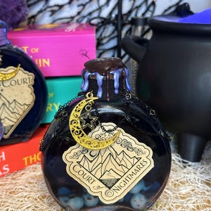 May include: A dark blue potion bottle with a label reading "Court of Nightmares." The bottle has a gold crescent moon and star charm, a dark brown stopper, and blue drips. The bottle contains blue orbs.