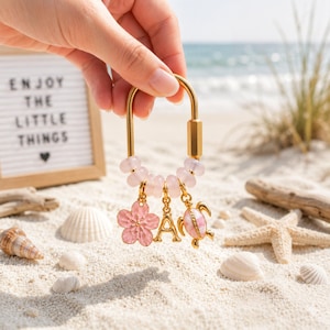 May include: A gold-toned U-shaped keyring featuring a row of pale pink beads and three dangling charms: a pink flower, the letter A, and a pink sea turtle. The keyring is held against a beach background with a sign reading ENJOY THE LITTLE THINGS.