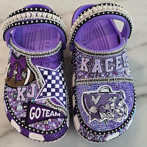 May include: Purple and white personalized Crocs with rhinestones and various embellishments, including a football, a checkered flag, a megaphone, a bow, and the text "Kacey", "KJ", and "Go Team".
