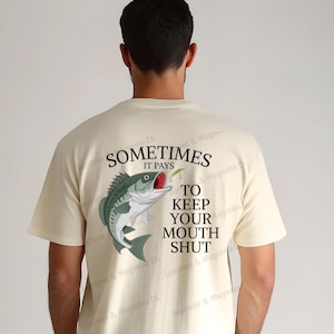 May include: Cream-colored t-shirt with a graphic of a fish and the text "Sometimes it pays to keep your mouth shut." The fish is green, white, and red. The shirt is short-sleeved and has a crew neck.