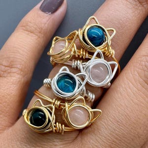 May include: A set of four gold wire wrapped rings with cat-shaped settings. Each ring features a different gemstone: rose quartz, turquoise, and two different shades of green.