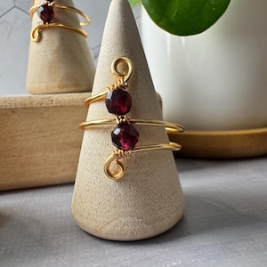 May include: Gold ring with two dark red gemstones wrapped in gold wire. The ring is displayed on a wooden ring holder.