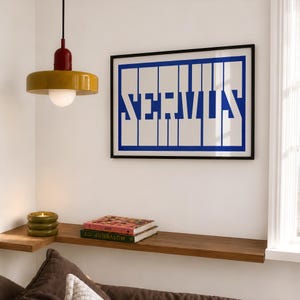May include: A rectangular wall art print featuring the word SERVUS in bold blue block letters against a white background, framed in black. A mustard yellow and red pendant light hangs nearby, and a stack of books sits on a wooden shelf.