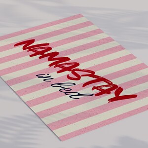 Framed poster with pink and cream vertical stripes, featuring the phrase “Namastay in bed” in bold red and navy handwritten fonts, styled as cozy bedroom wall decor.