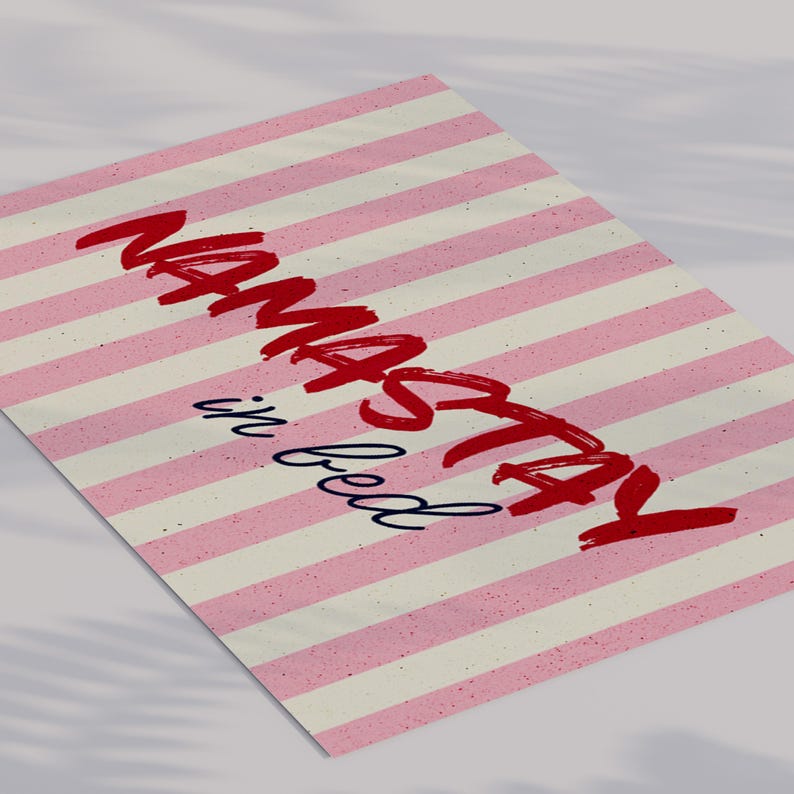Framed poster with pink and cream vertical stripes, featuring the phrase “Namastay in bed” in bold red and navy handwritten fonts, styled as cozy bedroom wall decor.