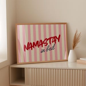Framed poster with pink and cream vertical stripes, featuring the phrase “Namastay in bed” in bold red and navy handwritten fonts, styled as cozy bedroom wall decor.