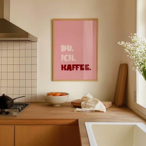 Framed poster with bold German text “Du. Ich. Kaffee.” in playful retro fonts on a solid pink background, leaning against a white wall.
