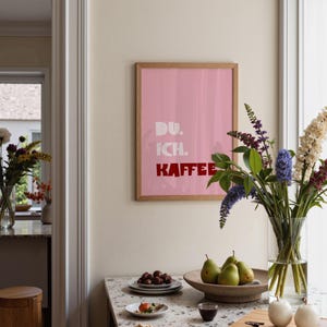 Framed poster with bold German text “Du. Ich. Kaffee.” in playful retro fonts on a solid pink background, leaning against a white wall.
