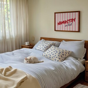 Framed poster with pink and cream vertical stripes, featuring the phrase “Namastay in bed” in bold red and navy handwritten fonts, styled as cozy bedroom wall decor.