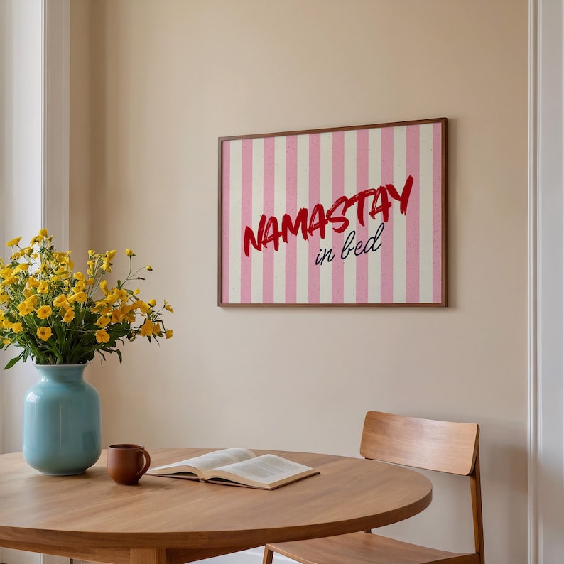 Framed poster with pink and cream vertical stripes, featuring the phrase “Namastay in bed” in bold red and navy handwritten fonts, styled as cozy bedroom wall decor.