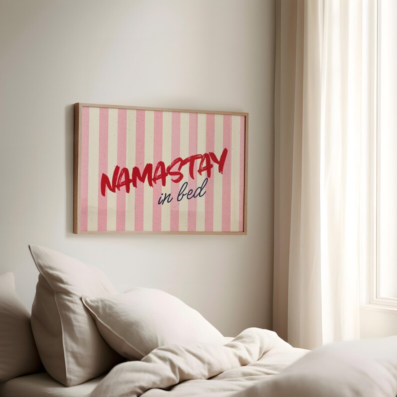 Framed poster with pink and cream vertical stripes, featuring the phrase “Namastay in bed” in bold red and navy handwritten fonts, styled as cozy bedroom wall decor.