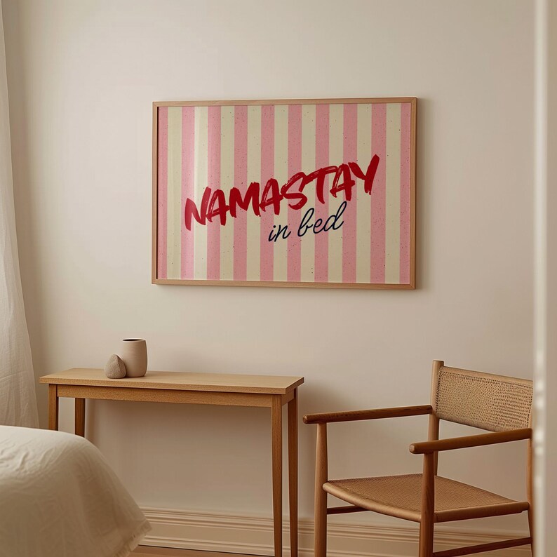 Framed poster with pink and cream vertical stripes, featuring the phrase “Namastay in bed” in bold red and navy handwritten fonts, styled as cozy bedroom wall decor.