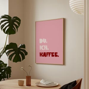 Framed poster with bold German text “Du. Ich. Kaffee.” in playful retro fonts on a solid pink background, leaning against a white wall.
