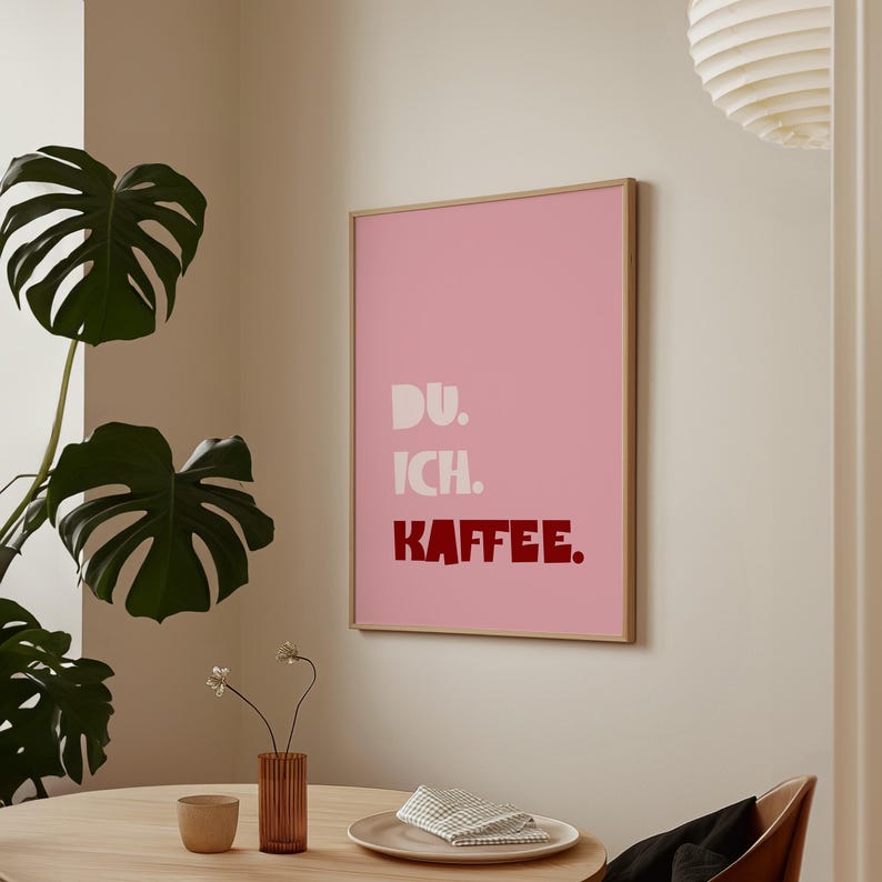 Framed poster with bold German text “Du. Ich. Kaffee.” in playful retro fonts on a solid pink background, leaning against a white wall.