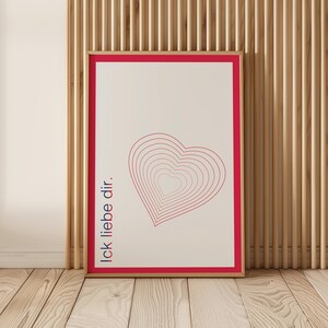 Peut inclure: Impression d'art encadrée avec un motif de cœur rouge composé de contours concentriques sur fond blanc cassé. L'expression "Ick liebe dir" est imprimée verticalement en rouge. L'œuvre est encadrée dans un cadre en bois avec un bord rouge, contre un mur en bois.