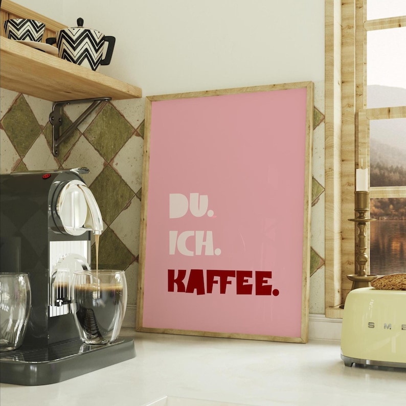 Framed poster with bold German text “Du. Ich. Kaffee.” in playful retro fonts on a solid pink background, leaning against a white wall.