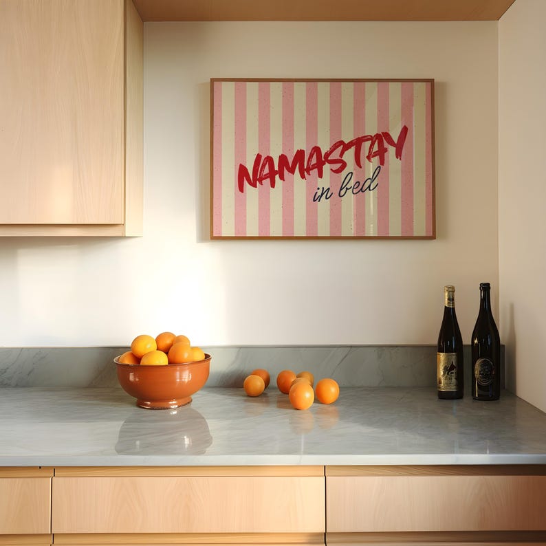 Framed poster with pink and cream vertical stripes, featuring the phrase “Namastay in bed” in bold red and navy handwritten fonts, styled as cozy bedroom wall decor.
