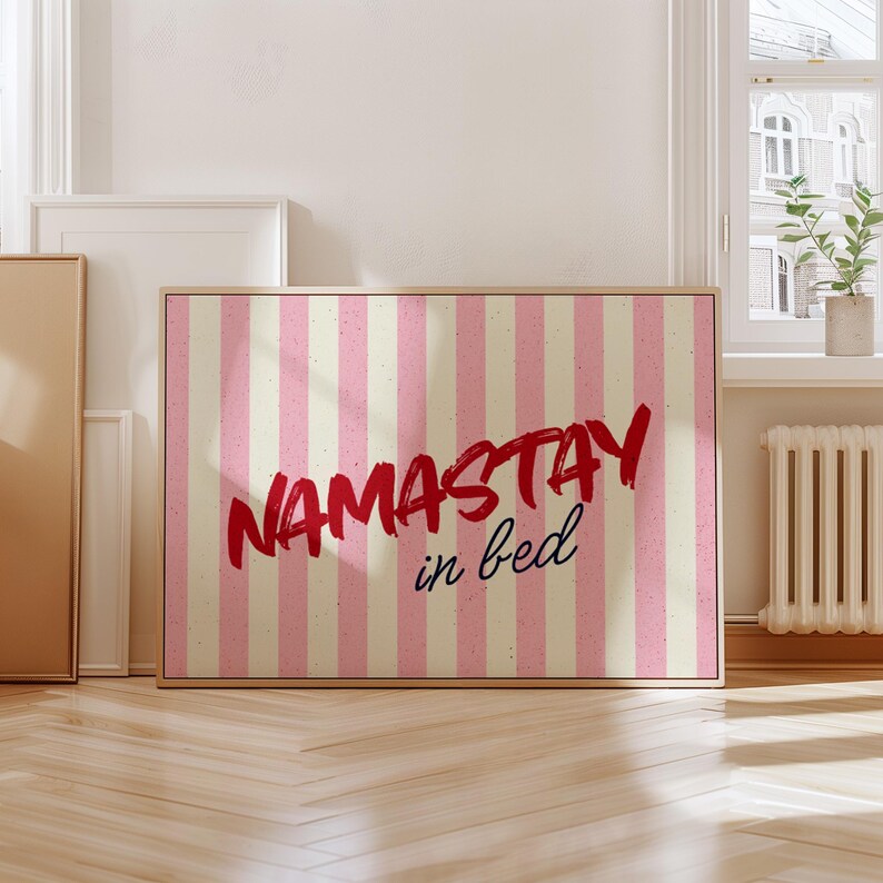 Framed poster with pink and cream vertical stripes, featuring the phrase “Namastay in bed” in bold red and navy handwritten fonts, styled as cozy bedroom wall decor.