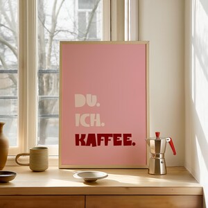 Framed poster with bold German text “Du. Ich. Kaffee.” in playful retro fonts on a solid pink background, leaning against a white wall.