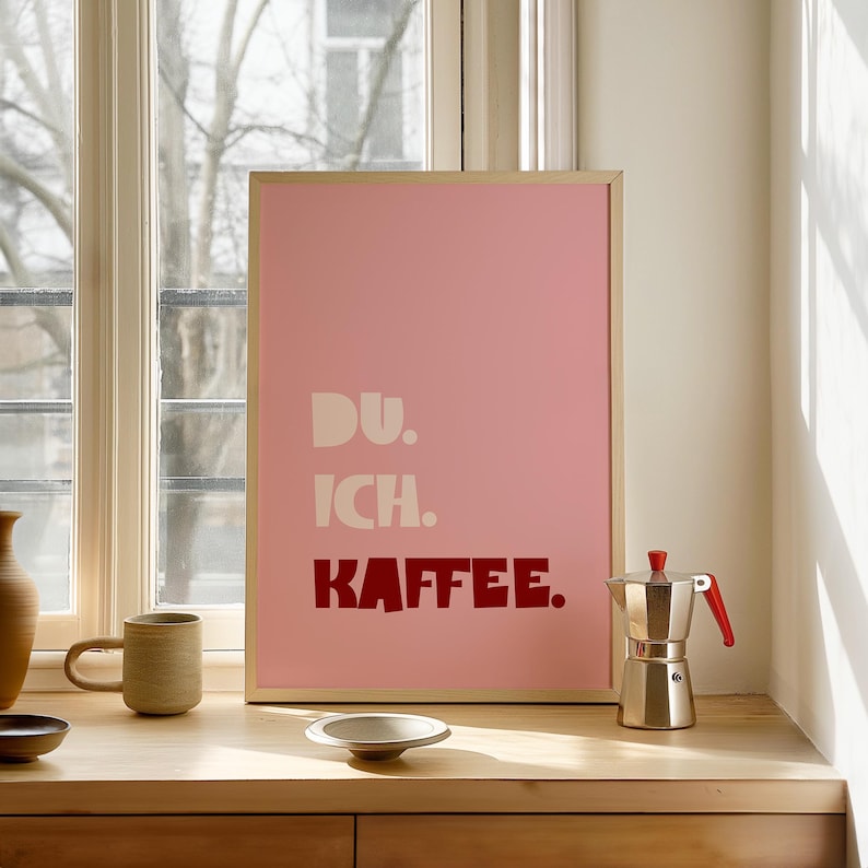 Framed poster with bold German text “Du. Ich. Kaffee.” in playful retro fonts on a solid pink background, leaning against a white wall.
