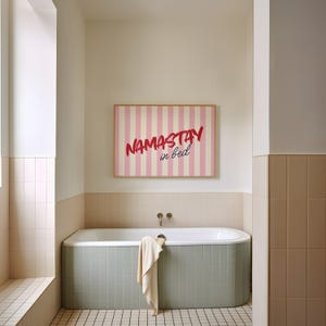 Framed poster with pink and cream vertical stripes, featuring the phrase “Namastay in bed” in bold red and navy handwritten fonts, styled as cozy bedroom wall decor.