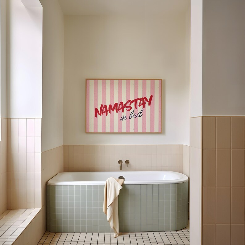 Framed poster with pink and cream vertical stripes, featuring the phrase “Namastay in bed” in bold red and navy handwritten fonts, styled as cozy bedroom wall decor.