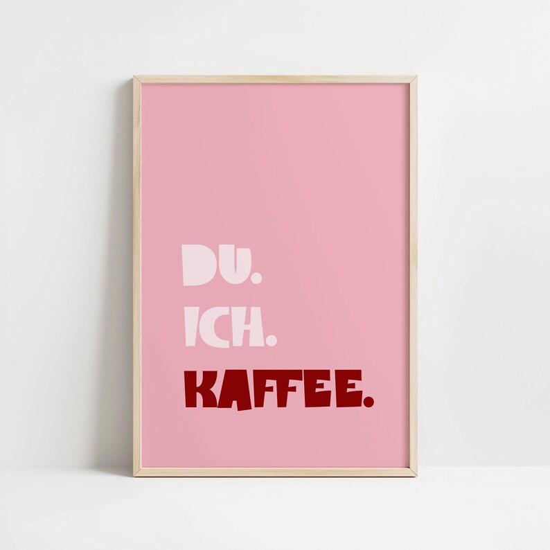 Framed poster with bold German text “Du. Ich. Kaffee.” in playful retro fonts on a solid pink background, leaning against a white wall.